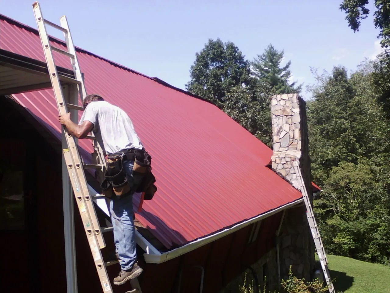 Roofer on a ladder beside a red metal roof and stone chimney — Soffit & Fascia Repair by Roofing Brothers in Port Jefferson
